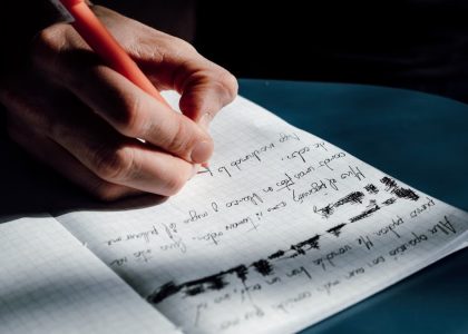 A person's hand writing in a notebook on a sunny day, showing detailed cursive notes.